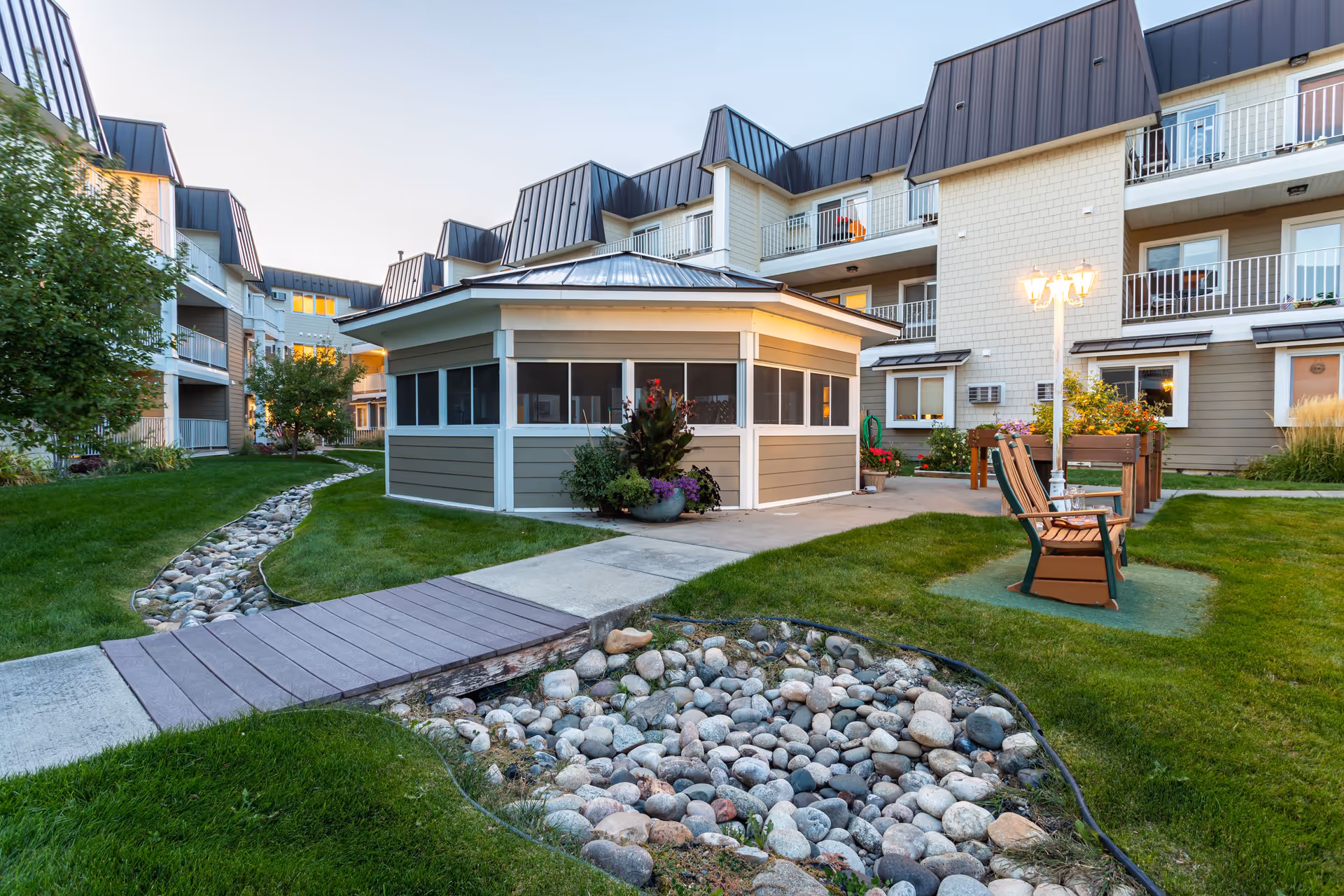 Outdoor courtyard area of Westpark Village senior living facility featuring a small wooden bridge over a dry rock bed, green grass, a gazebo-like structure with windows, wooden chairs near a lamp post, and multi-story residential buildings with balconies in the background.