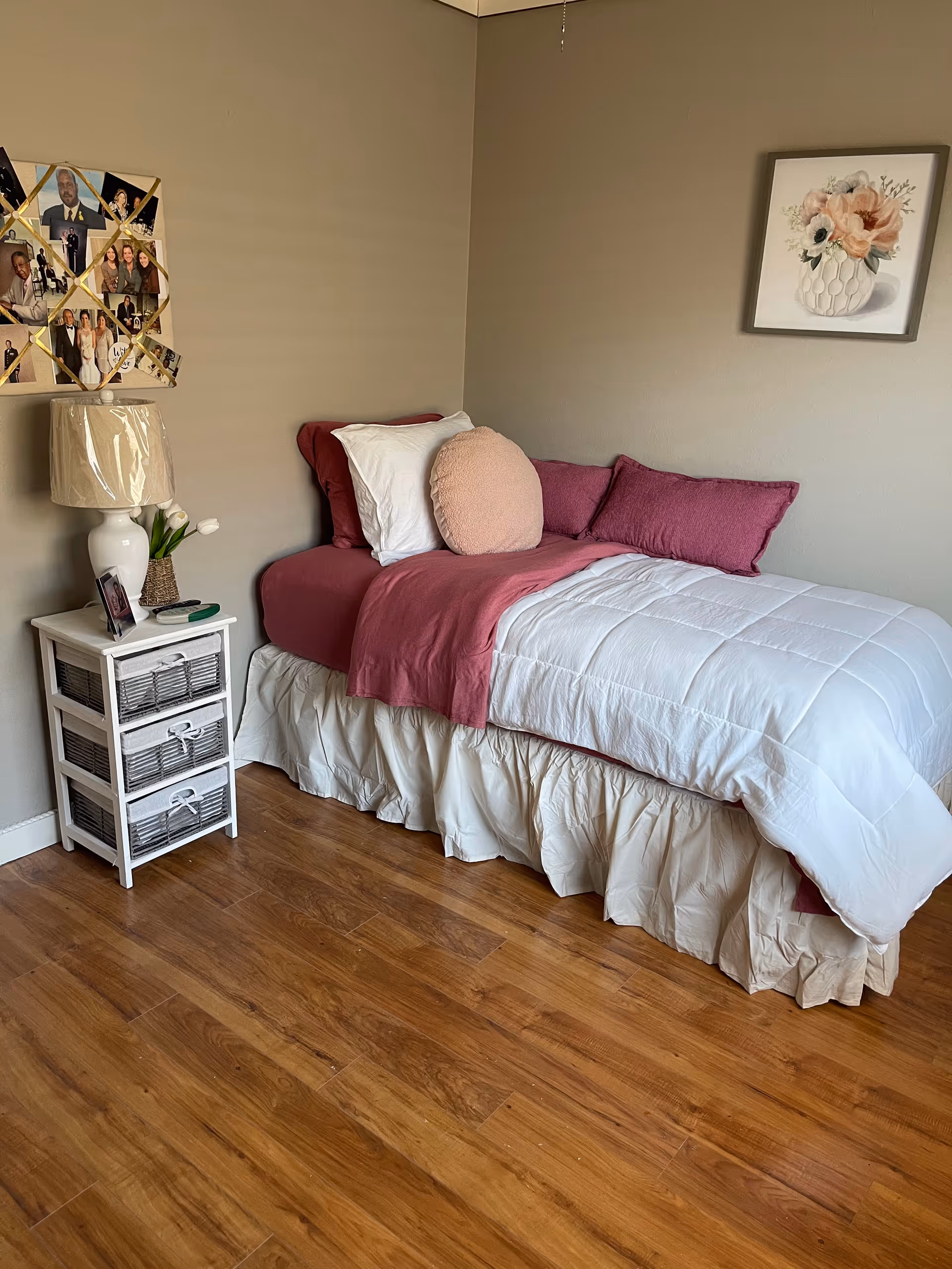 Neatly made single bed with white and pink bedding against a beige wall, a small white bedside table with a lamp and photo collage on the wall, and wood flooring.
