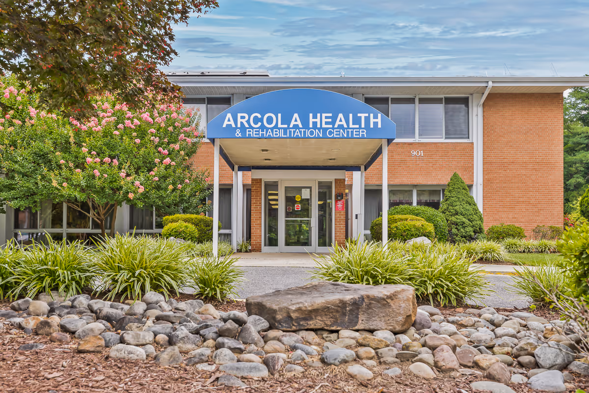 Front exterior view of Arcola Health & Rehabilitation Center building with a blue awning over the entrance, surrounded by landscaping including bushes, trees with pink flowers, and rocks in the foreground.