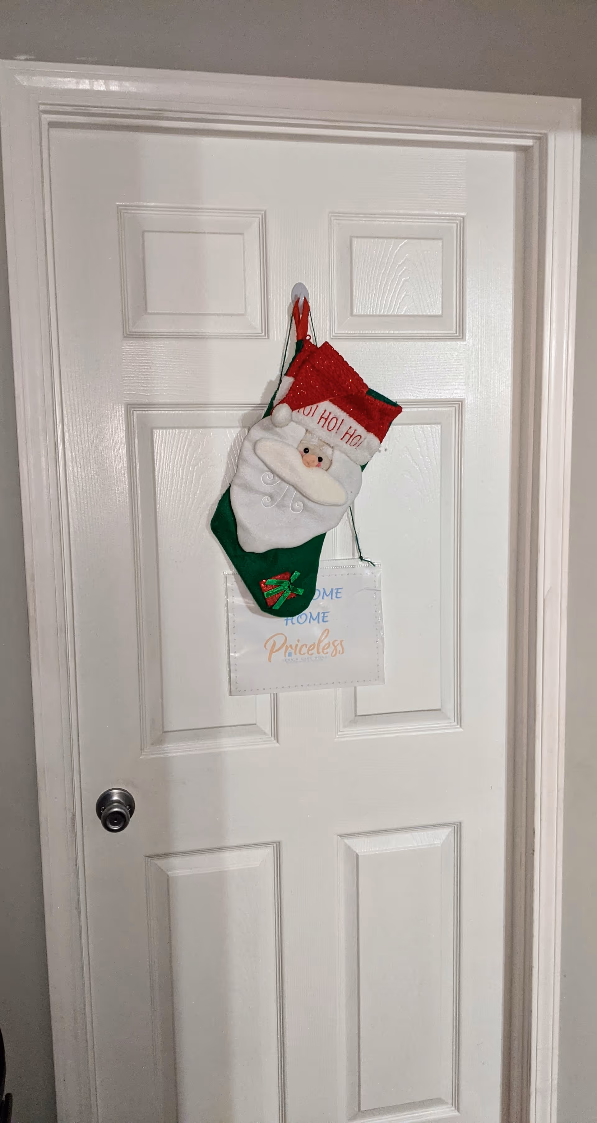 A white paneled door with a silver doorknob. Hanging on the door is a Christmas stocking featuring a Santa Claus face with the words 'HO! HO! HO!' on the red cuff. Below the stocking, there is a small sign that reads 'HOME HOME Priceless'.