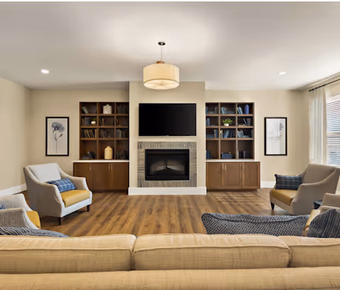 A cozy living room with a beige sofa in the foreground, two armchairs with yellow cushions, a fireplace with a mounted flat-screen TV above it, and built-in wooden bookshelves on either side. The room has wooden flooring, neutral-colored walls, framed artwork, and a ceiling light fixture.