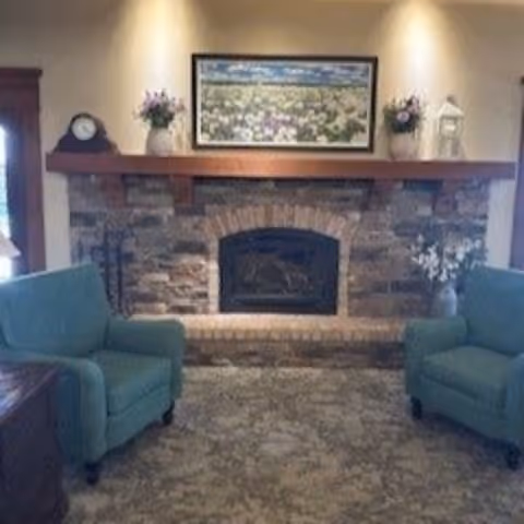 Cozy sitting area with a stone fireplace, wooden mantel holding a framed landscape and floral arrangements, flanked by two green armchairs.