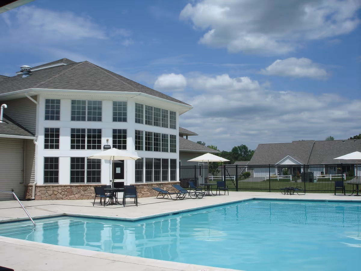 Outdoor swimming pool area with clear blue water, surrounded by a concrete deck with tables, chairs, and umbrellas. In the background, there is a large building with many windows and a stone base, along with a fenced grassy area and additional buildings under a partly cloudy sky.