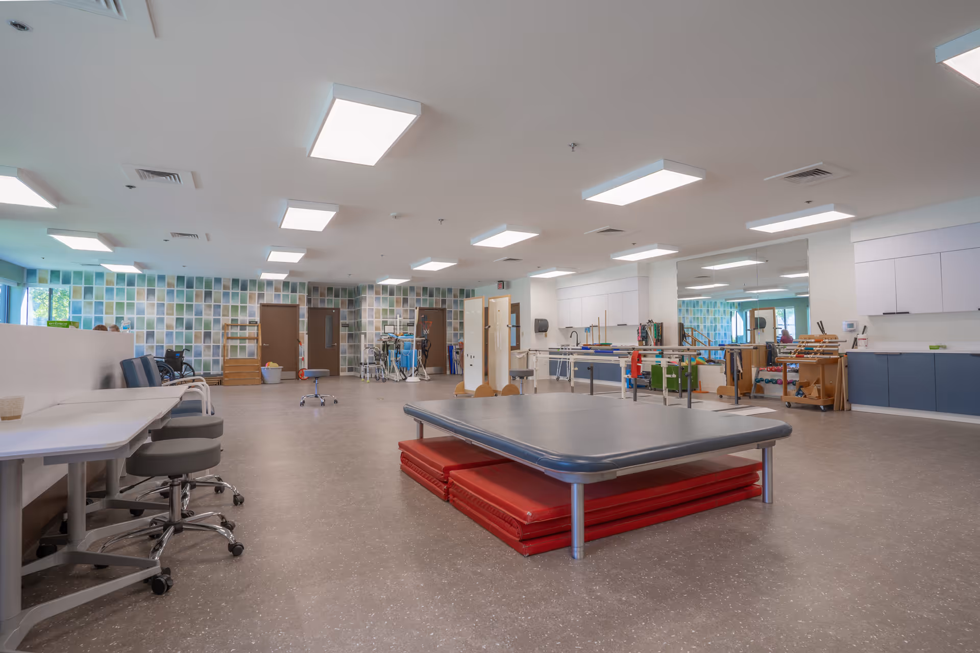 A spacious physical therapy room with exercise mats stacked under a large padded table in the center. The room has multiple chairs, tables, and various therapy equipment along the walls. The walls feature a colorful tiled pattern, and the ceiling has several rectangular fluorescent lights. There are cabinets and mirrors on one side of the room.