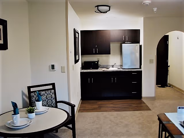 Interior view of a senior living facility unit showing a small kitchenette with dark cabinets, a mini refrigerator, microwave, and sink. In the foreground, there is a round dining table set with two place settings, napkins, and a small plant. The room has light-colored walls and carpet, with a wooden floor section in front of the kitchenette. An arched doorway leads to another area of the unit.