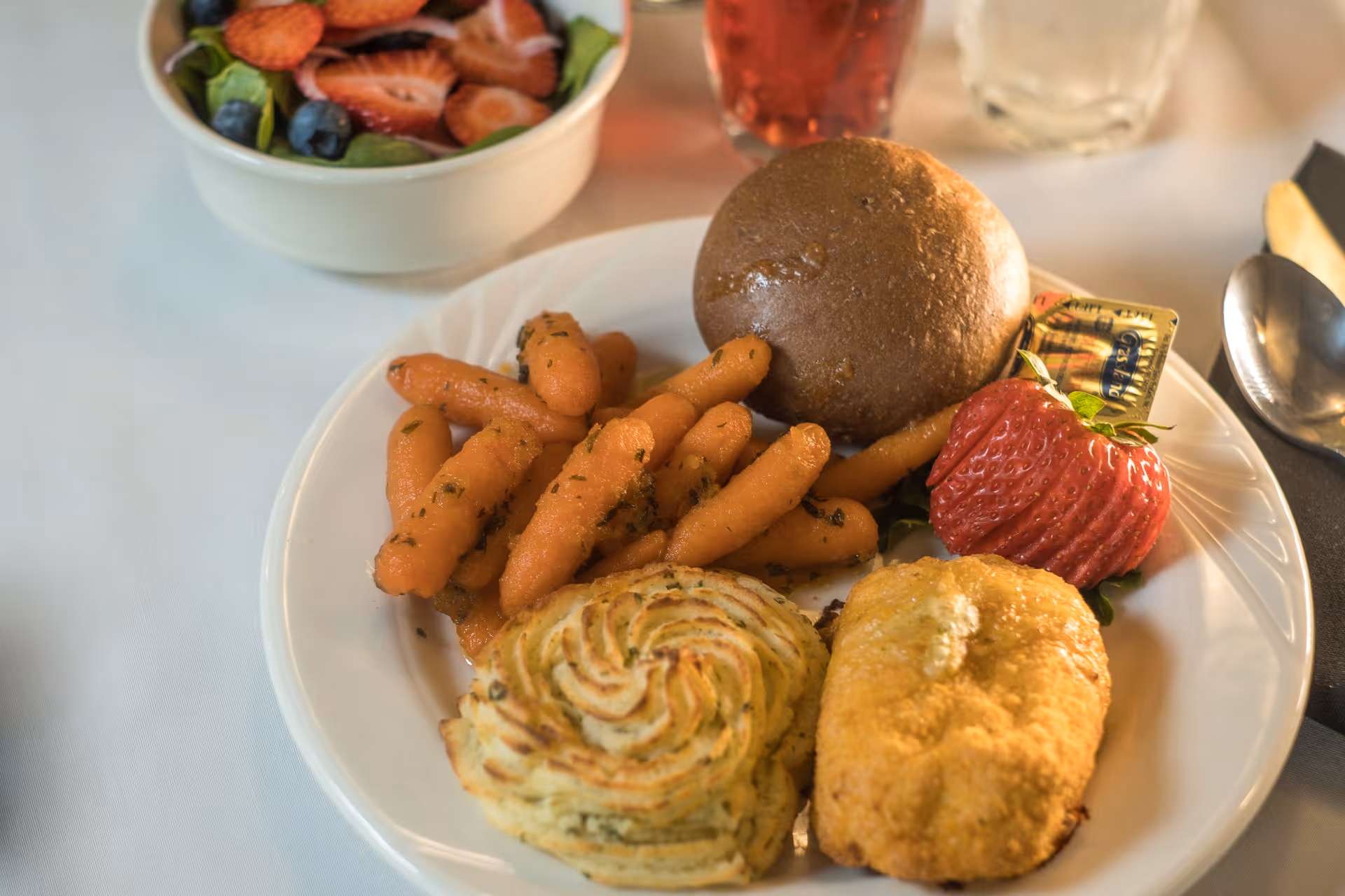 A plate of food featuring cooked baby carrots, a bread roll with a butter packet, a large strawberry, a serving of mashed potatoes piped in a decorative swirl, and a breaded fried item. In the background, there is a bowl of mixed fruit salad with strawberries and blueberries, and two glasses with beverages.