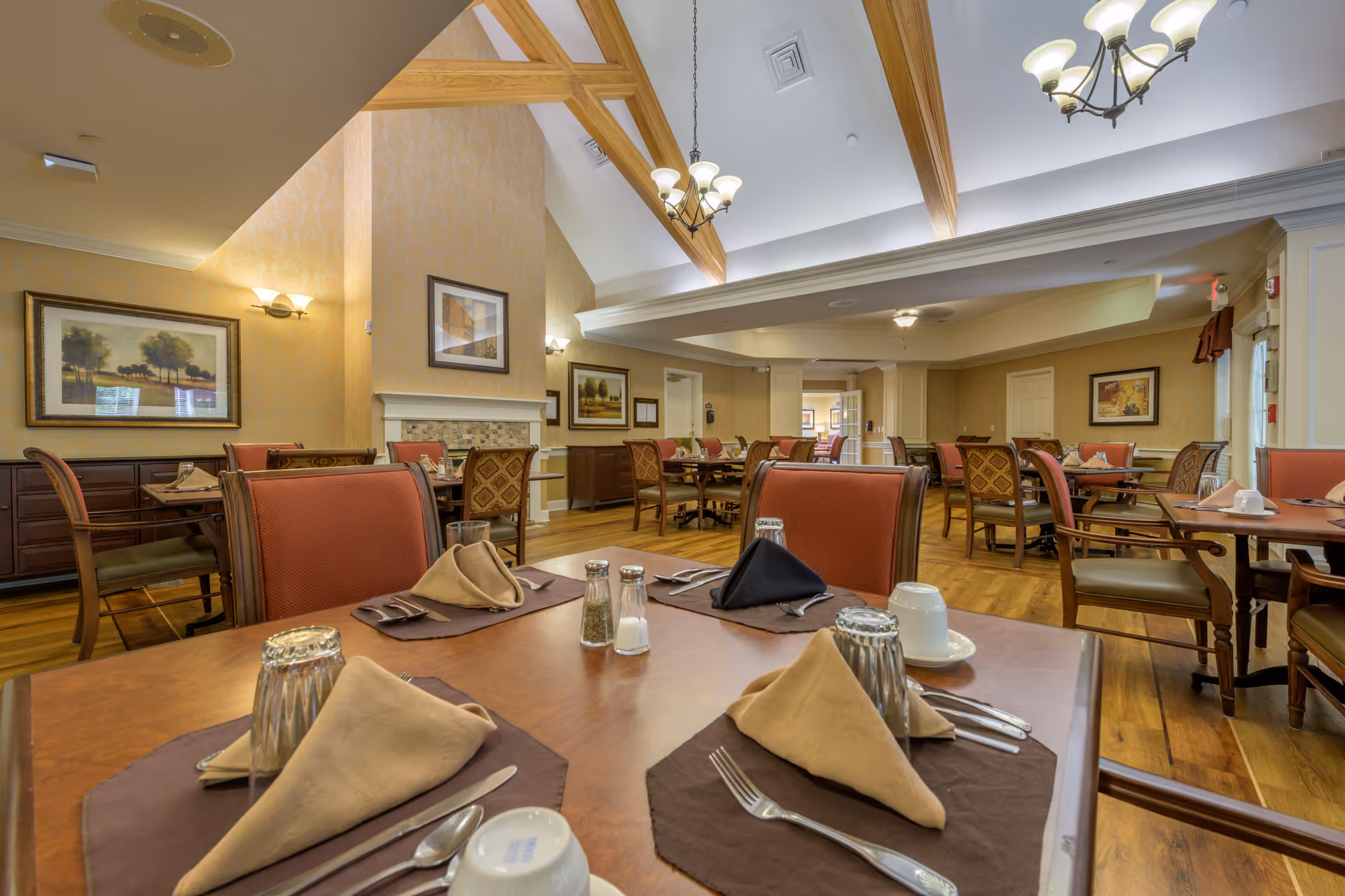 A spacious dining room with wooden floors and multiple tables set with brown placemats, folded napkins, silverware, salt and pepper shakers, and upside-down glasses. The room features high ceilings with wooden beams, chandeliers, framed artwork on the walls, and a fireplace. Chairs have red upholstery and wooden frames.