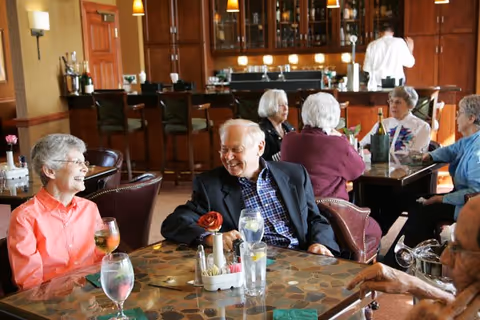A group of elderly people sitting and chatting around tables in a cozy dining area with a bar in the background. The atmosphere is warm and social, with drinks on the tables and a staff member attending behind the bar.
