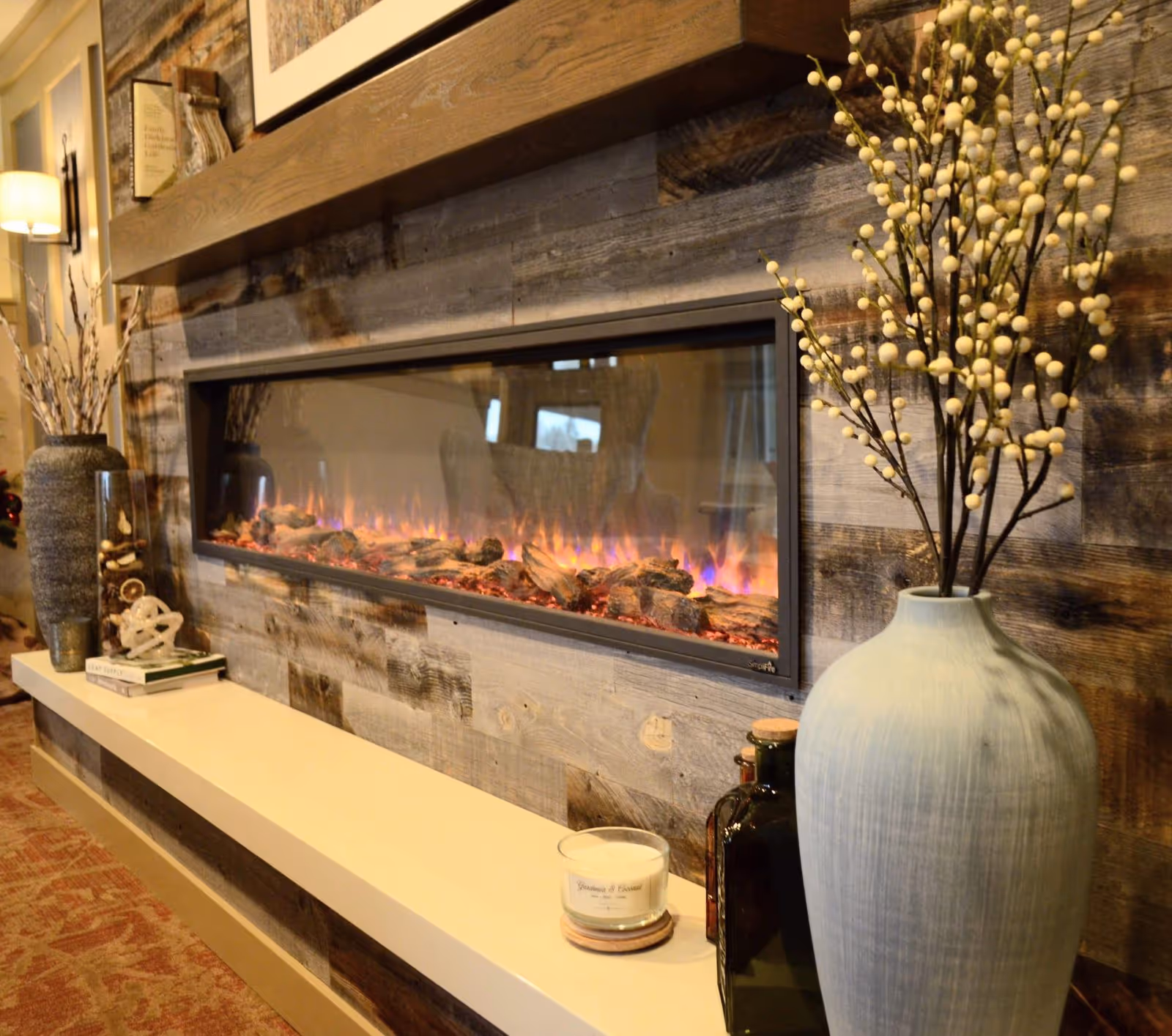 A cozy interior scene featuring a modern electric fireplace set into a rustic wooden wall. On the mantel above the fireplace are framed pictures and decorative items. Below the fireplace, a white shelf holds a candle, a large blue vase with white berry branches, and other decorative bottles. A lamp and additional decor are visible in the background.