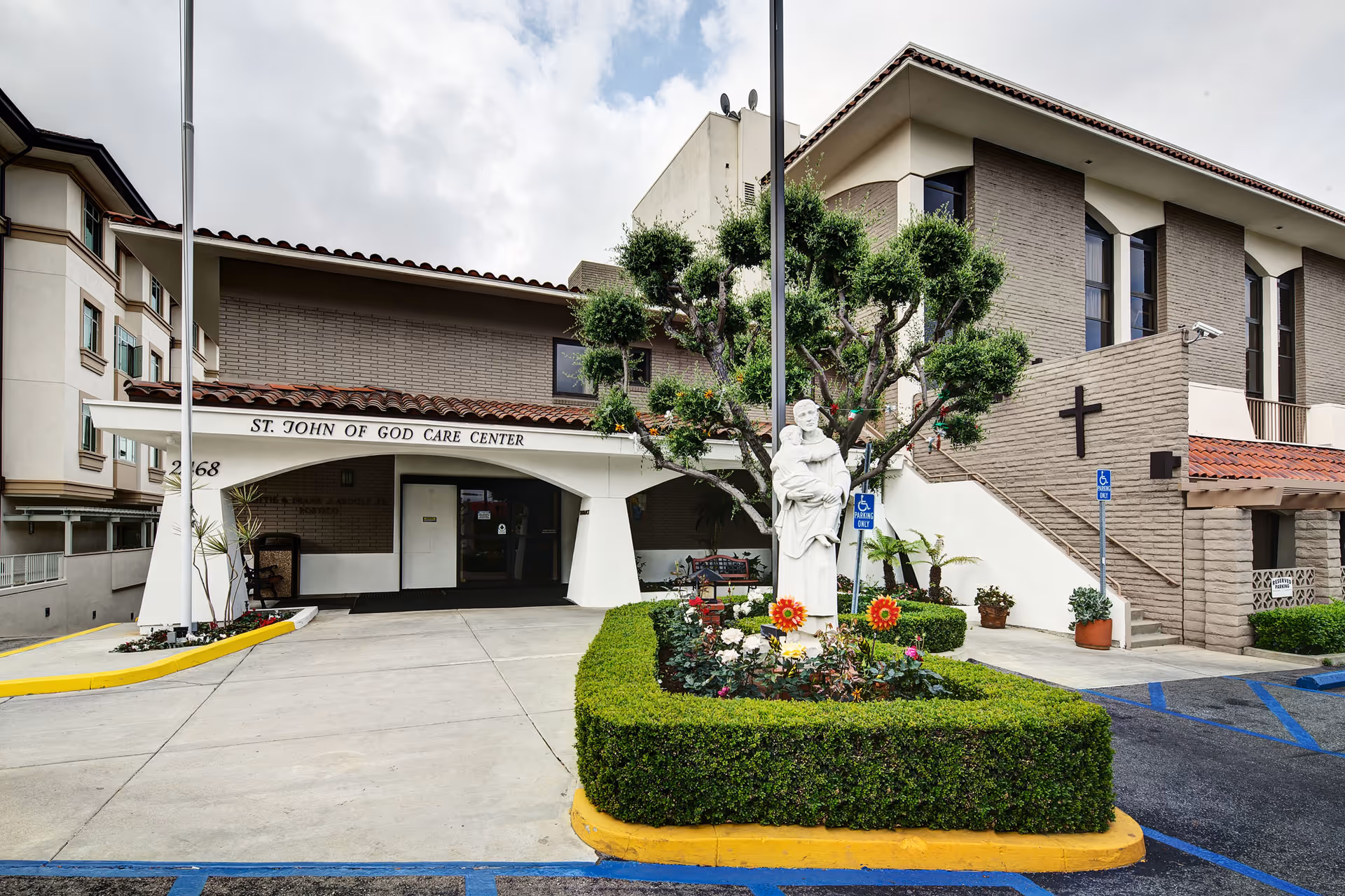 Exterior view of St. John of God Care Center showing the entrance with a covered walkway, a statue of a saint holding a child surrounded by a neatly trimmed hedge and flowers, a tree, and a building with beige brick walls and a cross mounted on one side. There are handicap parking signs and spaces in front of the building.