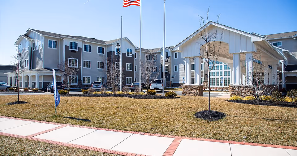 Front exterior of a senior living building with a covered entrance, flagpoles, lawn, and a sidewalk.