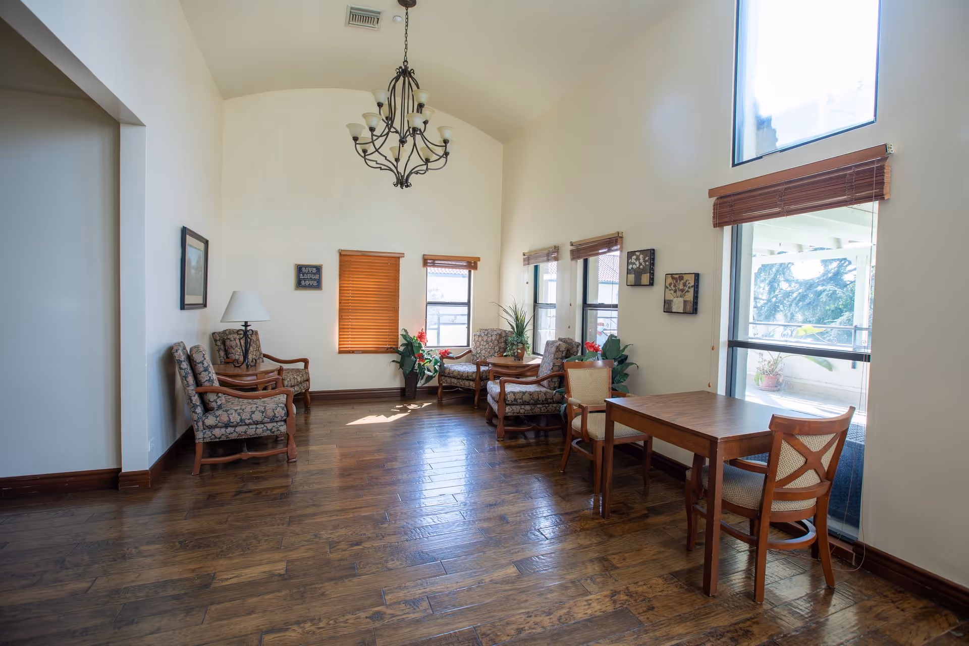 A bright and spacious living room area with wooden flooring, several upholstered armchairs with floral patterns, a wooden table with two chairs, potted plants, wall art, and large windows with wooden blinds letting in natural light. A decorative chandelier hangs from the ceiling.
