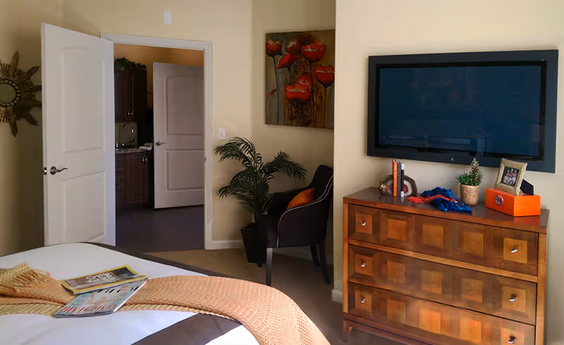 A furnished bedroom with a bed, wooden dresser topped by a wall-mounted TV, an armchair and an open doorway to another room.