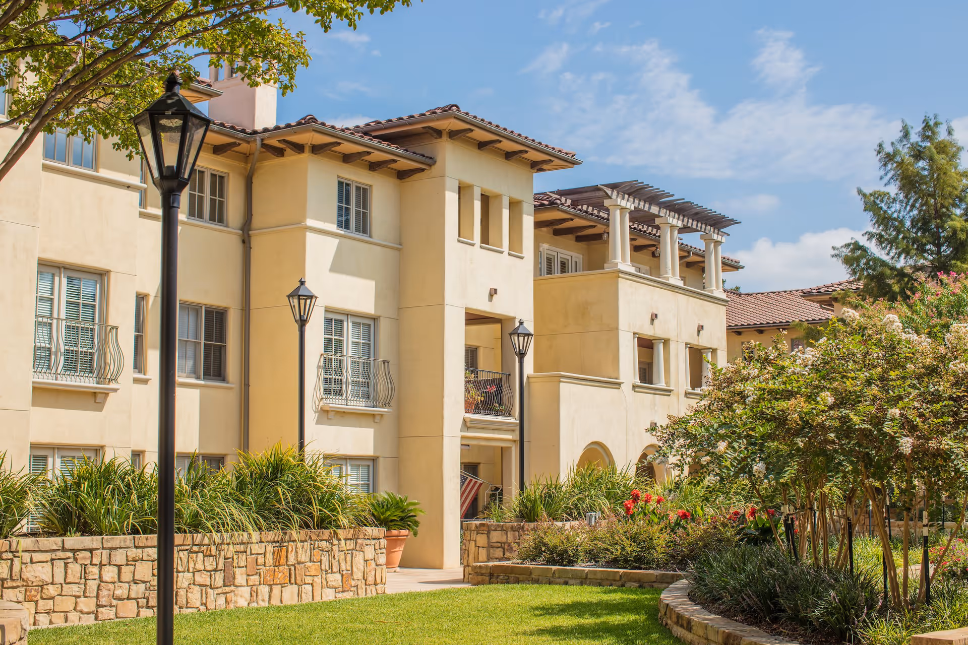 Exterior view of a beige multi-story senior living facility building with balconies, surrounded by well-maintained landscaping including green grass, bushes, and flowering plants under a partly cloudy sky.
