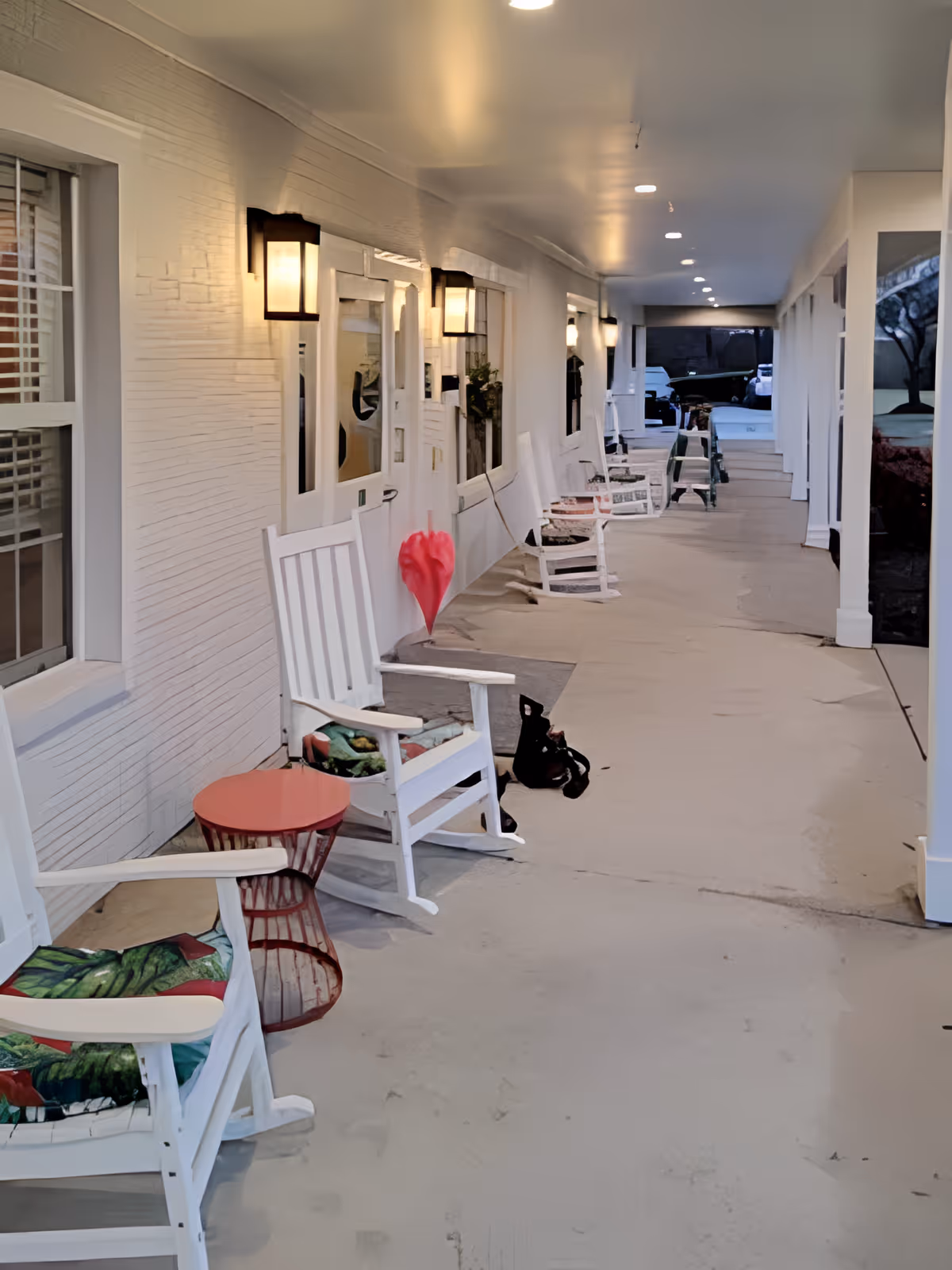 Covered outdoor walkway with white rocking chairs and small tables lined up against the wall of a building. Wall-mounted lights illuminate the area, and there are windows and doors along the wall. A red umbrella is leaning against the wall near one of the chairs.