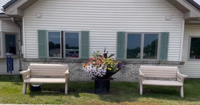 Exterior view of a single-story building with beige siding and green shutters on the windows. In front of the building, there are two beige benches on either side of a large black planter filled with colorful flowers. The area is grassy and well-maintained.