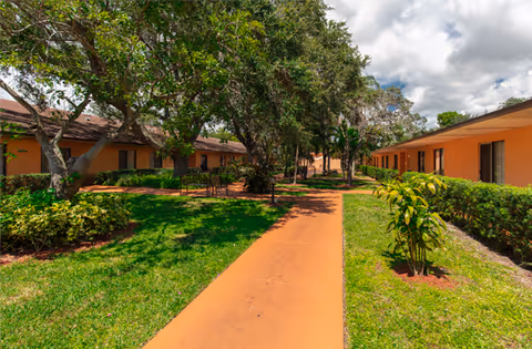 Outdoor view of a senior living facility with a paved walkway flanked by green lawns, bushes, and trees. Single-story buildings with orange walls and brown roofs line both sides of the walkway under a partly cloudy sky.