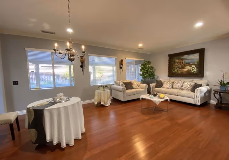A spacious living room with wooden flooring, two beige sofas with decorative pillows, a glass coffee table with a lace cloth and decorative items, a round table covered with a white tablecloth and a dark runner, a chandelier hanging from the ceiling, two windows with blinds, wall sconces, a large framed painting on the wall, and several potted plants.