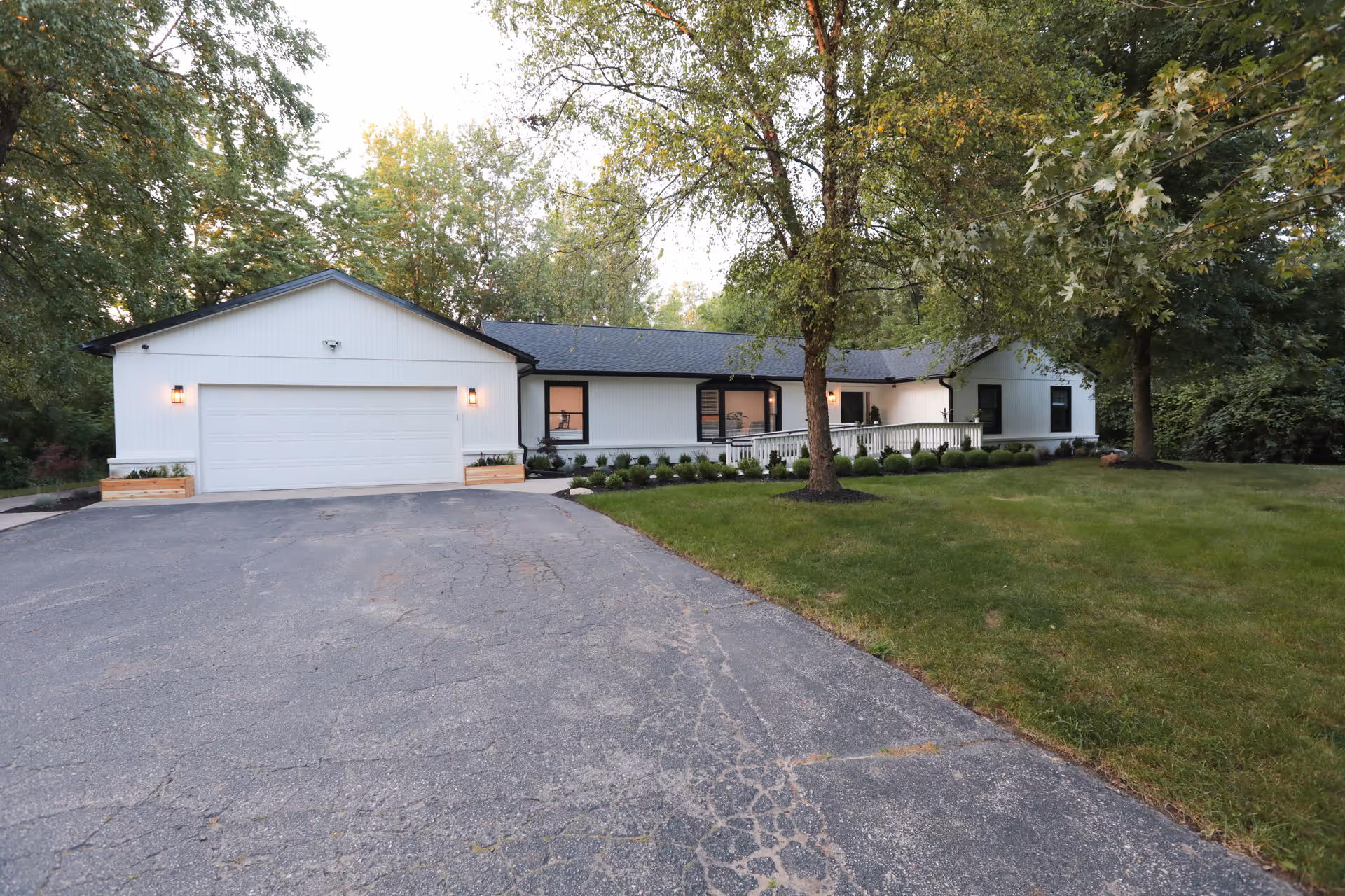Exterior view of a single-story white building with a dark roof, surrounded by trees and greenery. The building has a large garage door on the left and a ramp leading to the entrance on the right. The driveway and lawn are visible in the foreground.