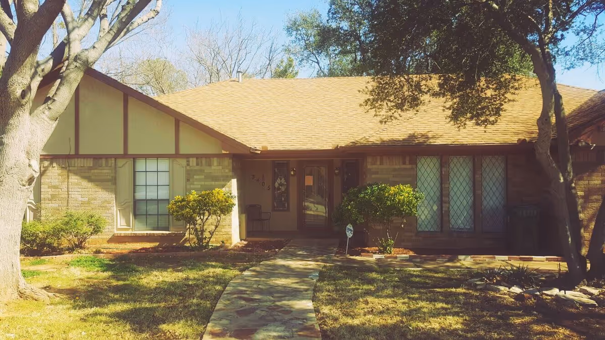 Front exterior view of a single-story brick house with a sloped roof, a stone pathway leading to the front door, and trees and bushes in the yard.