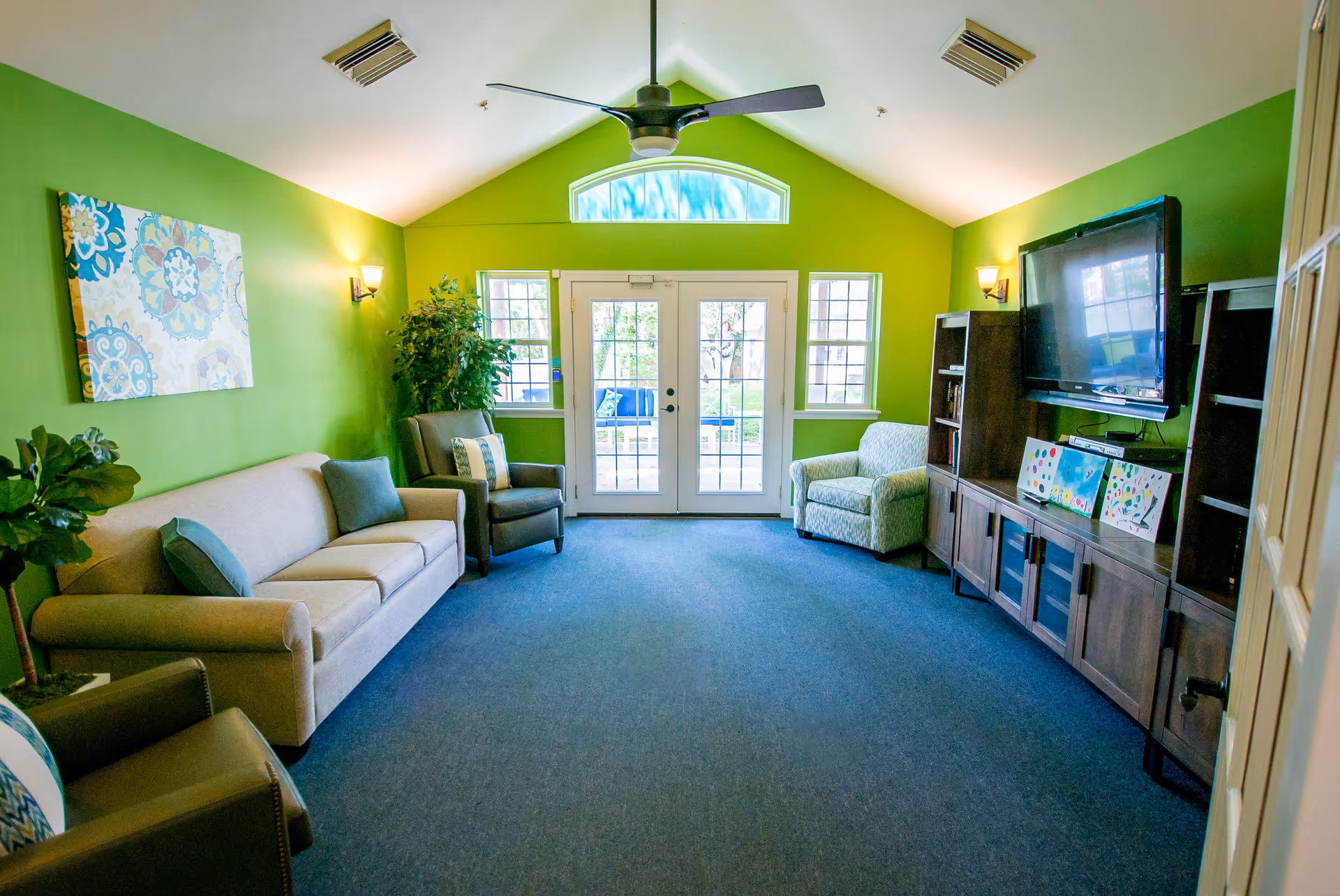 A bright living room with green walls, a beige sofa, two armchairs, a ceiling fan, a flat-screen TV mounted on a wooden cabinet, and French doors with windows letting in natural light.