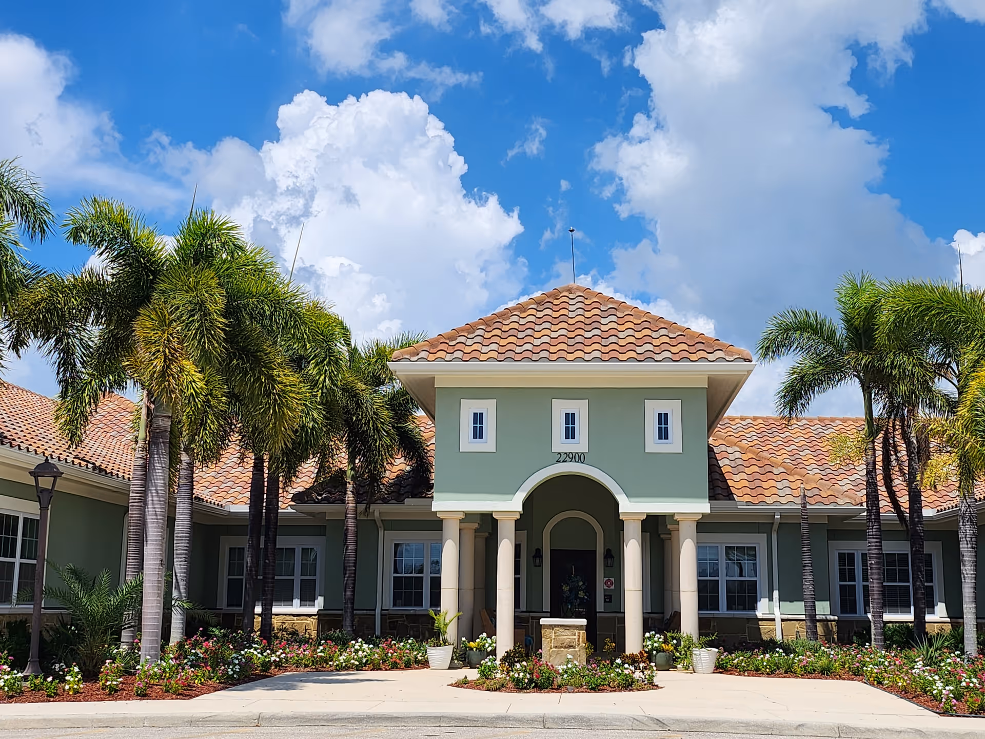 Front exterior view of Gulf Coast Memory Care facility with a green building, terracotta tiled roof, palm trees, and landscaped flower beds under a blue sky with clouds.