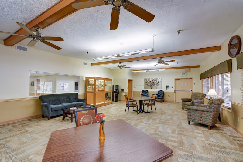 Spacious senior living common room with tables, chairs, sofas, ceiling fans, and windows.