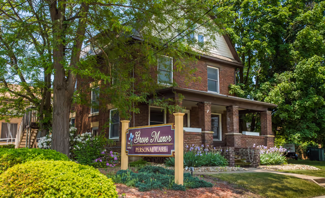 Exterior view of a two-story brick building with a covered porch, surrounded by green trees and shrubs. A sign in front reads 'Grove Manor Personal Care'.
