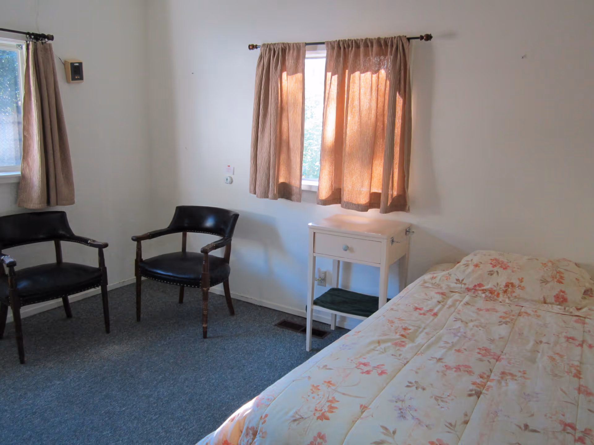 A simple bedroom with a single bed covered in a floral patterned comforter, two black wooden chairs with armrests, a small white nightstand with a drawer and a shelf, and two windows with brown curtains letting in natural light.