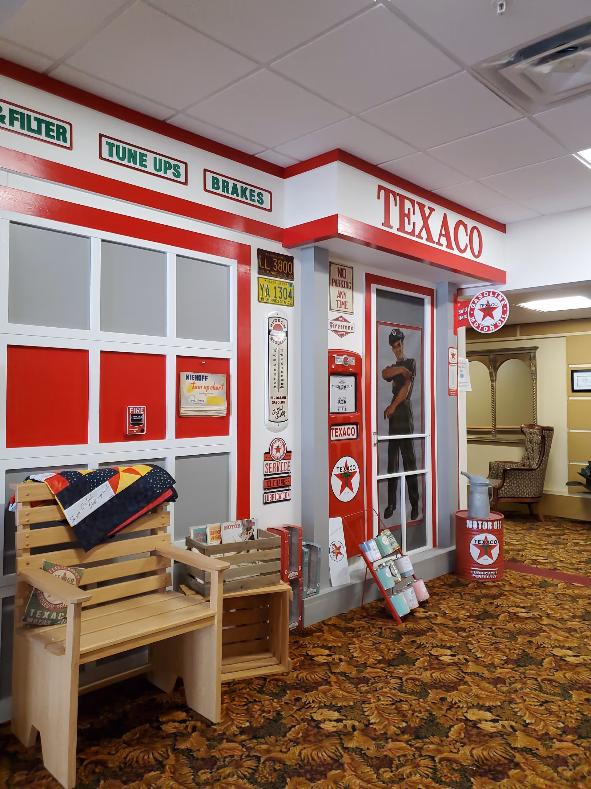 Interior hallway decorated with a vintage Texaco gas station theme, featuring red and white wall panels, old license plates, Texaco signs, a wooden bench with a quilt, and a patterned carpet. There is a painted figure of a gas station attendant on a door and various vintage oil cans and advertisements displayed.