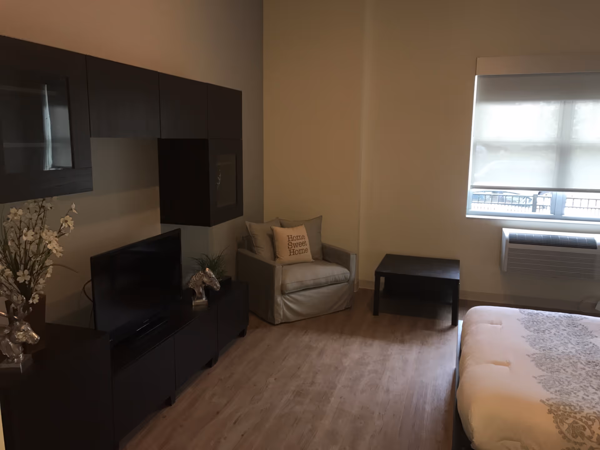 A cozy living space with a beige armchair featuring a 'Home Sweet Home' pillow, a small black coffee table, a bed with a patterned white and beige comforter, a flat-screen TV on a dark wooden cabinet, and a window with a white roller shade partially drawn. The room has light wood flooring and neutral-colored walls.
