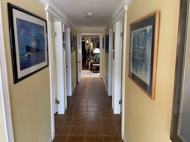 A narrow hallway with beige walls and brown tiled floor, lined with framed pictures on both sides. Several white doors are visible along the hallway, leading to other rooms. At the end of the hallway, a living area with furniture and a lamp is partially visible.