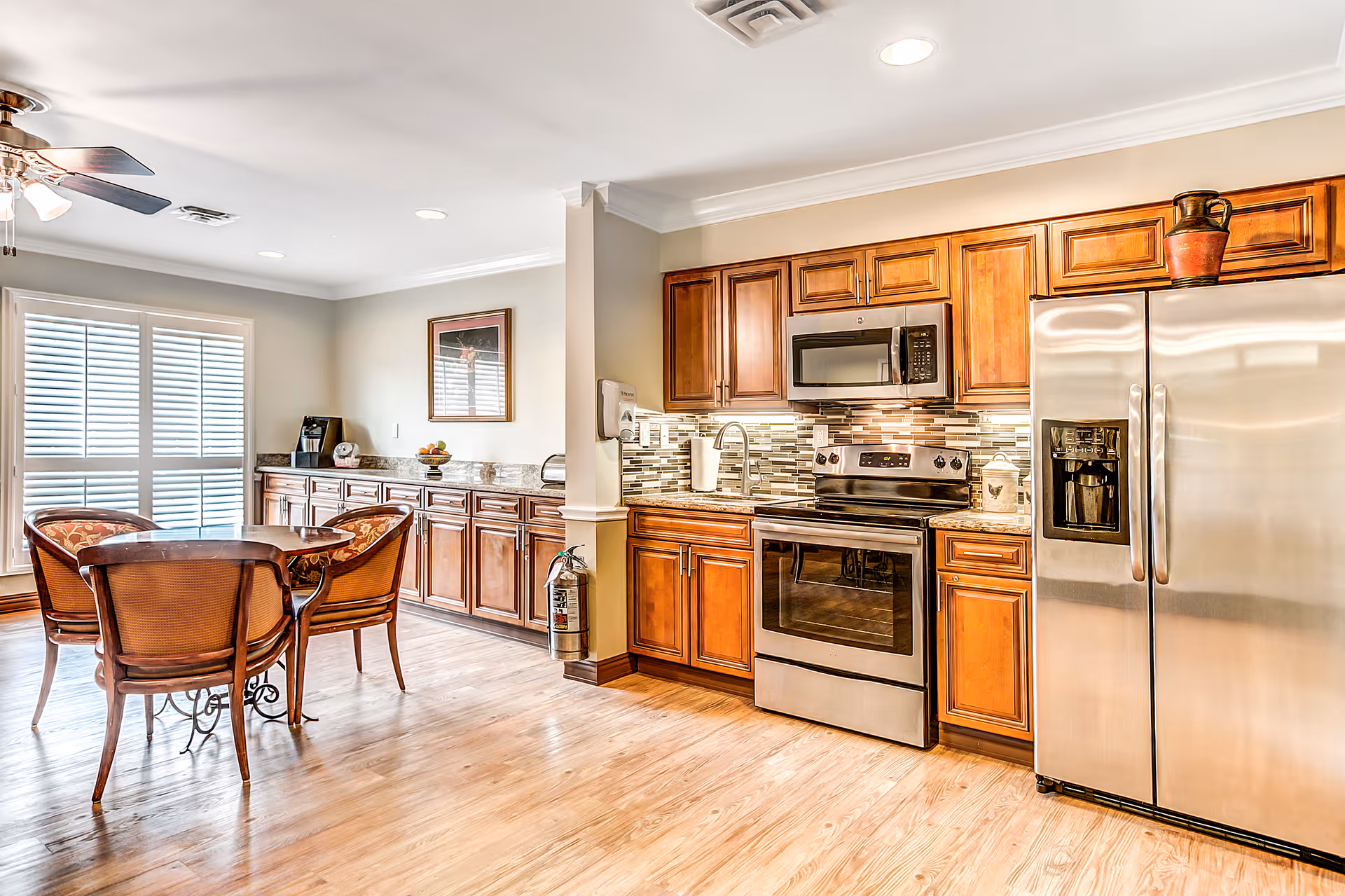 A bright kitchen and dining area with wooden cabinets, stainless steel appliances including a refrigerator, oven, and microwave. There is a small round dining table with four wooden chairs, a ceiling fan with lights, and a large window with white blinds letting in natural light. The floor is light wood, and there is a fire extinguisher mounted on the wall near the cabinets.