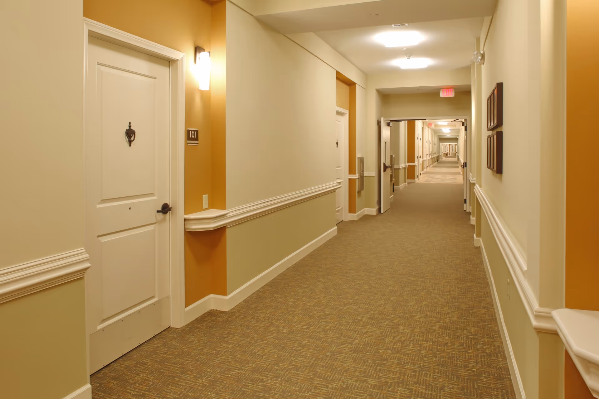 A long, well-lit hallway in a senior living facility with beige and light green walls, white doors with room numbers, and patterned carpet flooring. The hallway has wall-mounted lights and framed pictures on one side.