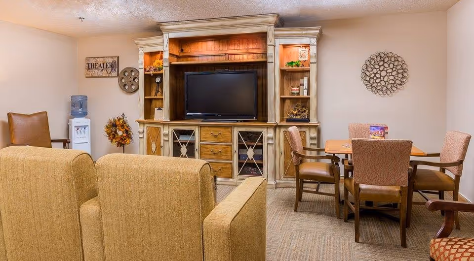 A cozy common area with a large wooden entertainment center holding a flat-screen TV, surrounded by shelves with decorative items. In front of the TV are two beige upholstered chairs. To the right, there is a wooden table with four cushioned chairs. On the left side, a water cooler and a decorative wreath are visible, along with a wall sign that reads 'THEATER'. The room has neutral-colored walls and carpeted flooring.