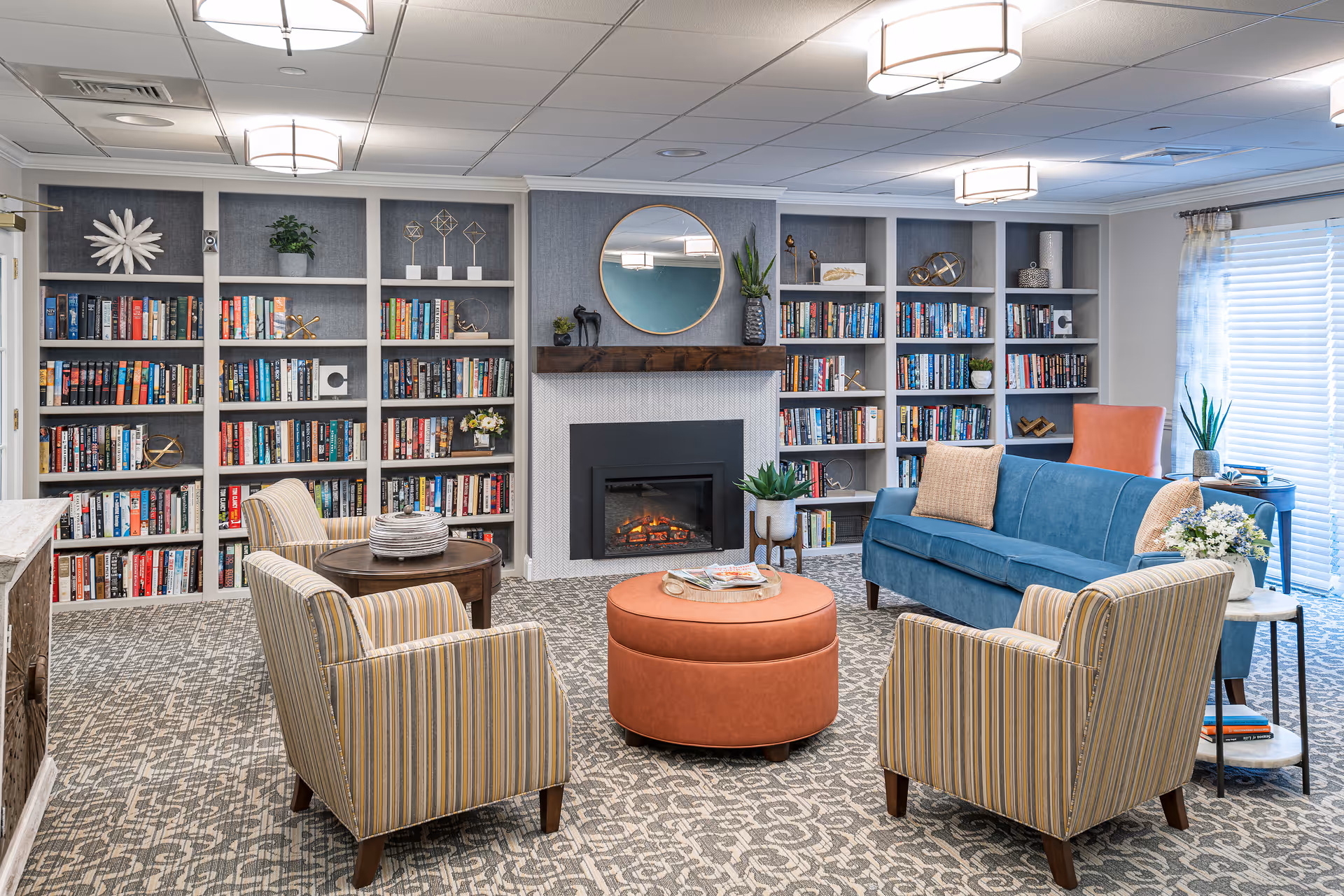A cozy living room with a blue sofa, two striped armchairs, and a round orange ottoman in the center. Behind the seating area is a fireplace with a round mirror above it, flanked by built-in bookshelves filled with books and decorative items. The room has patterned carpet flooring, ceiling lights, and a window with blinds and sheer curtains on the right side.