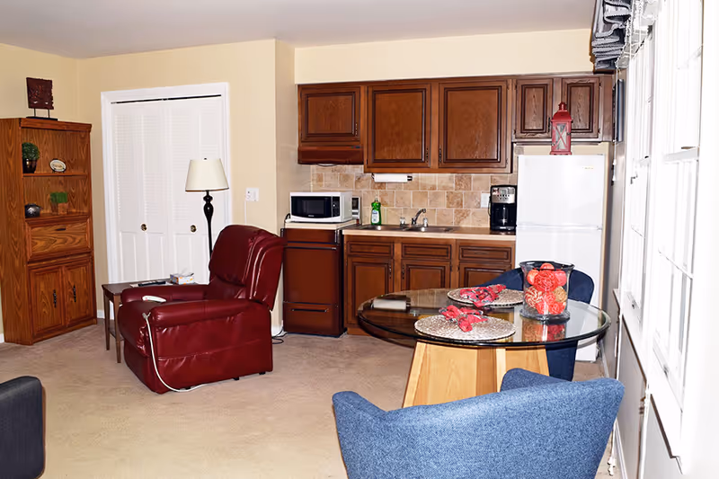 A cozy living area with a small kitchen in the background. The kitchen has wooden cabinets, a white refrigerator, a microwave, a coffee maker, and a sink. In the foreground, there is a round glass table with two blue chairs and a decorative centerpiece. To the left, there is a red recliner chair next to a wooden bookshelf and a floor lamp. The room has beige carpet and light-colored walls with windows on the right side.