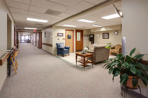 A long, well-lit hallway in a senior living facility with beige walls and carpeted floors. On the right side, there is a small seating area with a blue armchair, a patterned armchair, a wooden coffee table, and a beige chair. The walls have framed pictures and a clock. A large potted plant is visible in the foreground on the right.