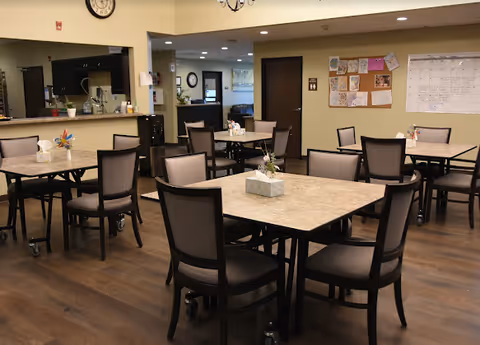Communal dining room with multiple square tables and chairs, a serving counter, and a bulletin board in the background.