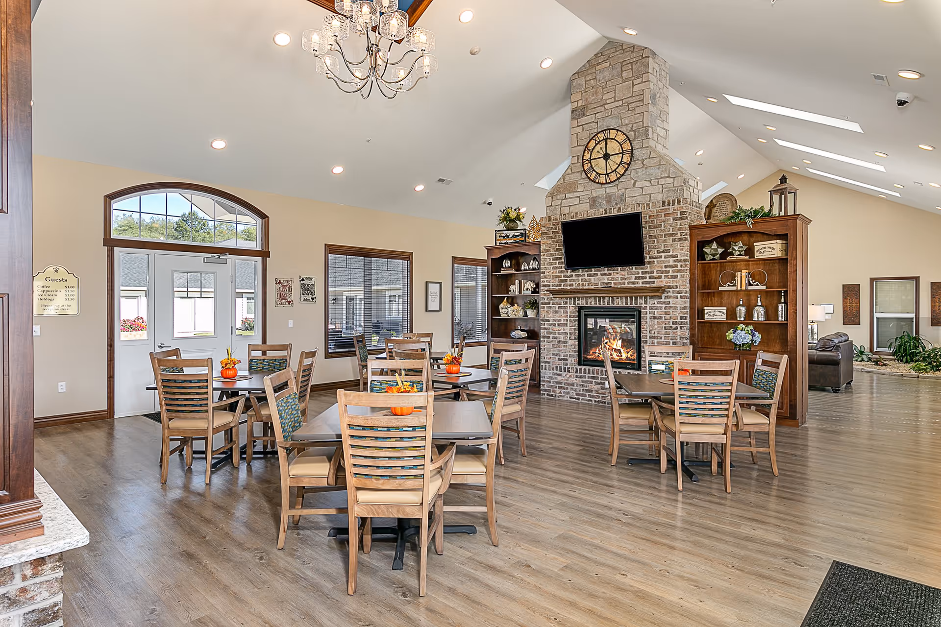 A bright and spacious dining area with wooden tables and chairs arranged neatly. The room features a large brick fireplace with a mounted TV above it, flanked by wooden shelves decorated with plants and ornaments. There are large windows letting in natural light, a chandelier hanging from the ceiling, and a door with a transom window above it. The floor is wooden, and the overall ambiance is warm and inviting.