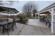 Outdoor patio area with several tables and chairs, some under a large umbrella, adjacent to a covered pavilion with additional seating. Leafless trees and a fenced grassy area are visible in the background under an overcast sky.