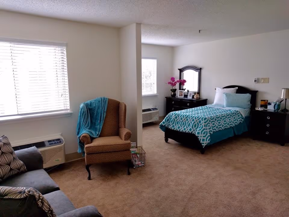 A cozy bedroom with a single bed covered in a blue and white patterned bedspread, a wooden dresser with a mirror and decorative items including a vase with pink flowers, a nightstand with a lamp and clock, a comfortable armchair with a blue throw blanket, and a sofa with patterned cushions. The room has beige carpet, white walls, and two windows with blinds allowing natural light to enter.
