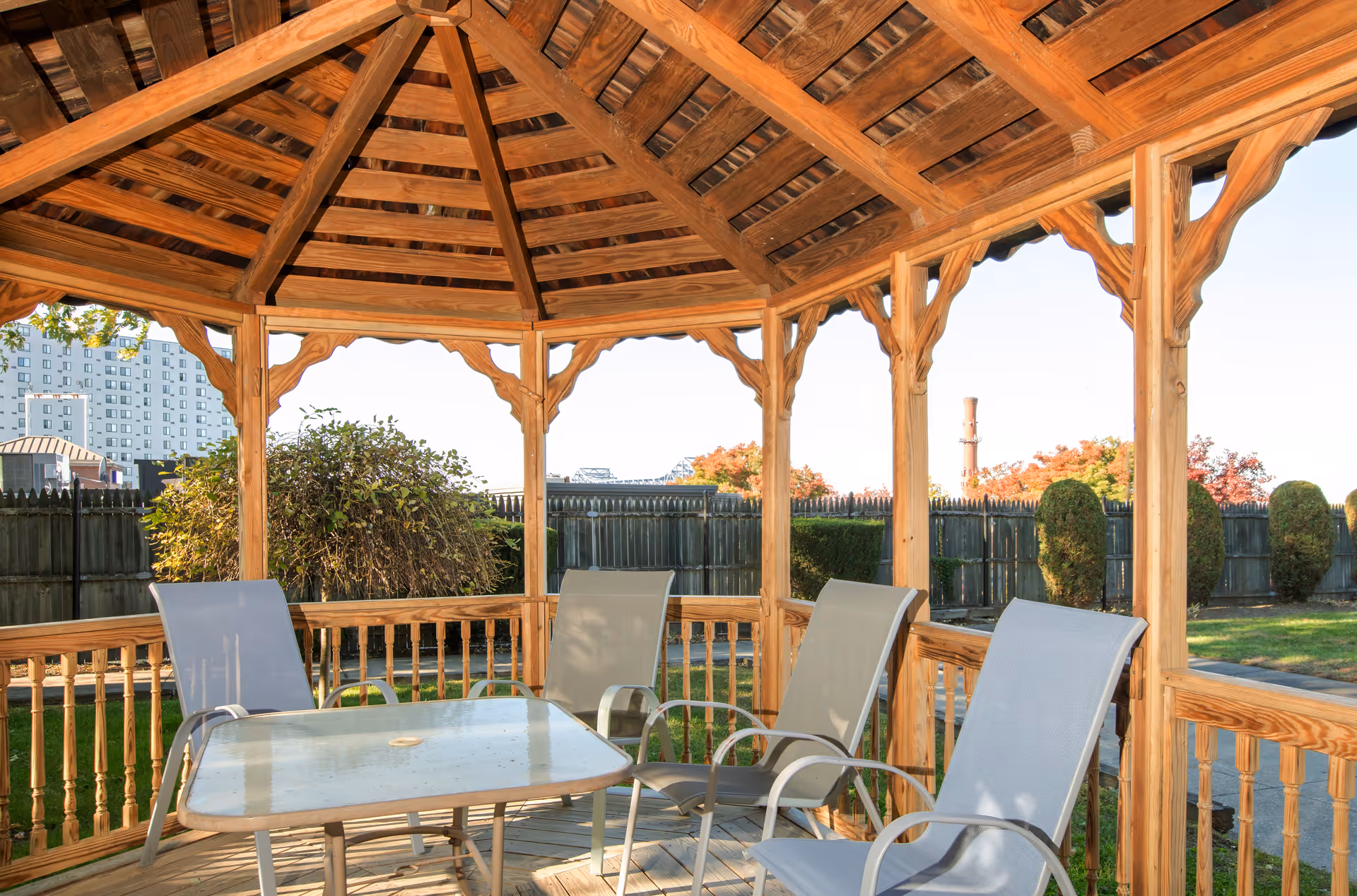 Wooden gazebo with a glass-top table and four outdoor chairs inside, situated in a fenced garden area with trimmed bushes and trees visible in the background under a clear sky.