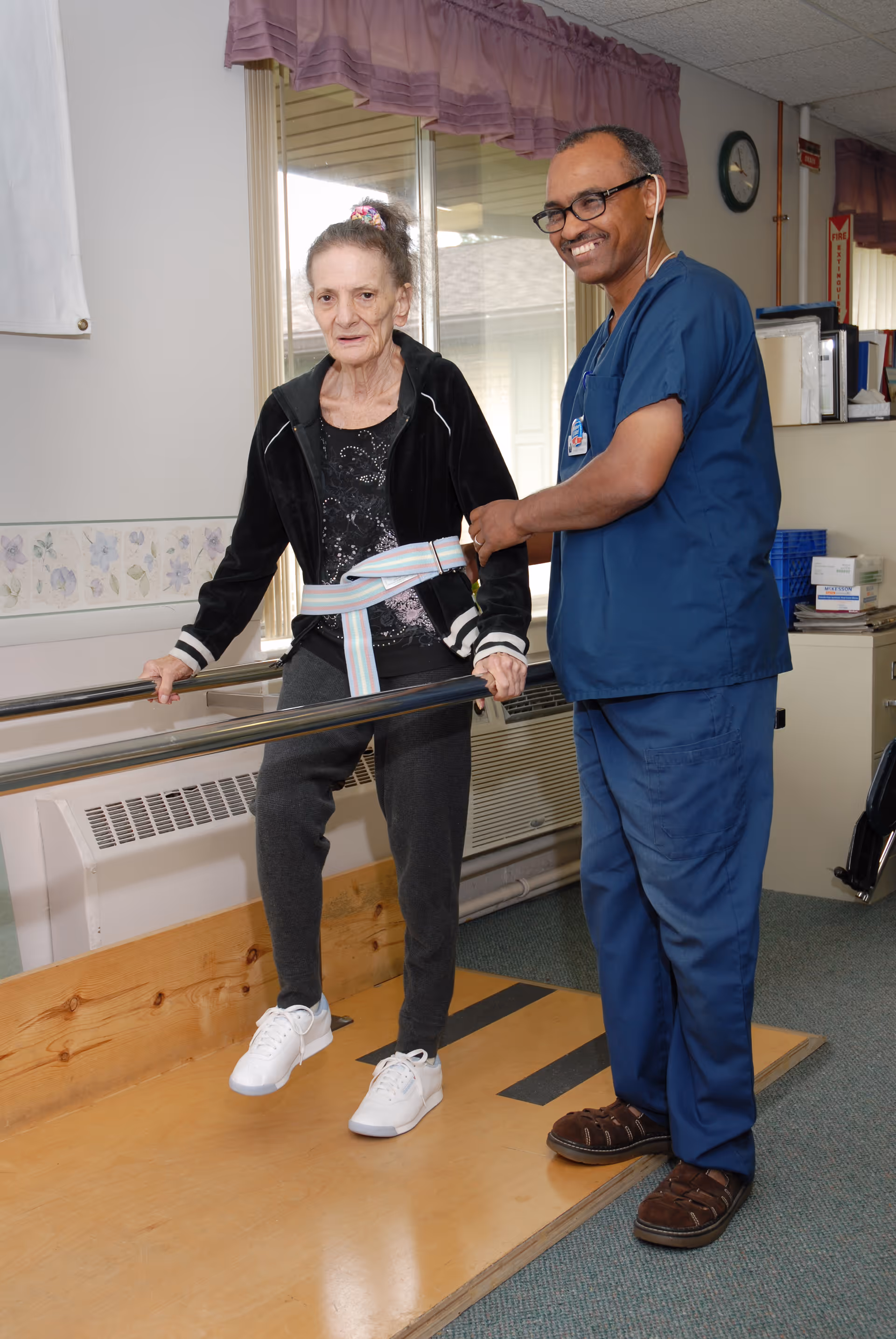 An elderly woman wearing a black jacket and gray pants is using parallel bars for walking assistance while a healthcare professional in blue scrubs supports her with a gait belt in a rehabilitation setting.