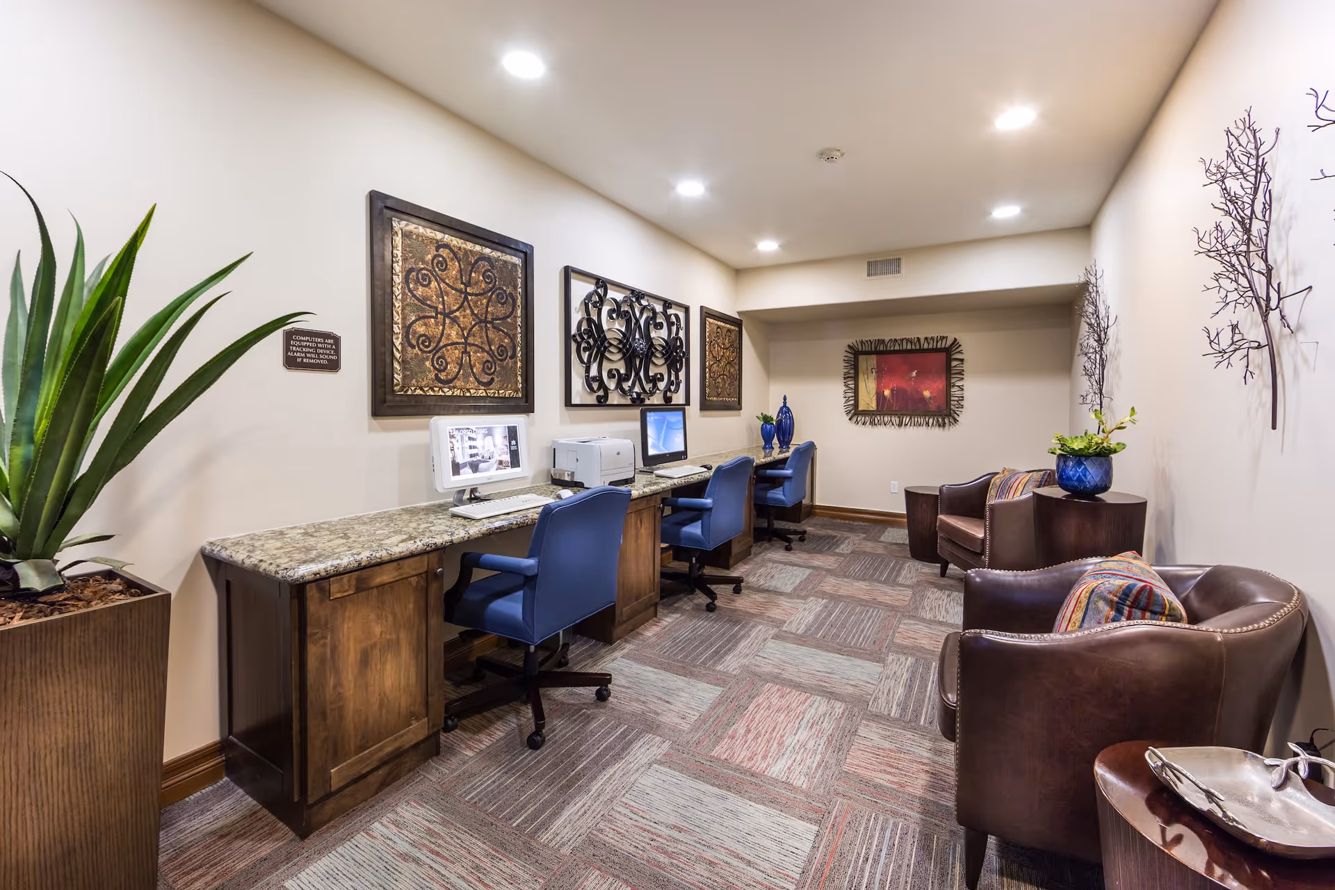 A cozy computer room in a senior living facility with three computer stations along a granite countertop, each with a blue office chair. The walls are decorated with framed metal and fabric artwork. There are two brown leather armchairs with colorful cushions and small wooden tables with plants and decorative items. The room has patterned carpet tiles and recessed ceiling lights.