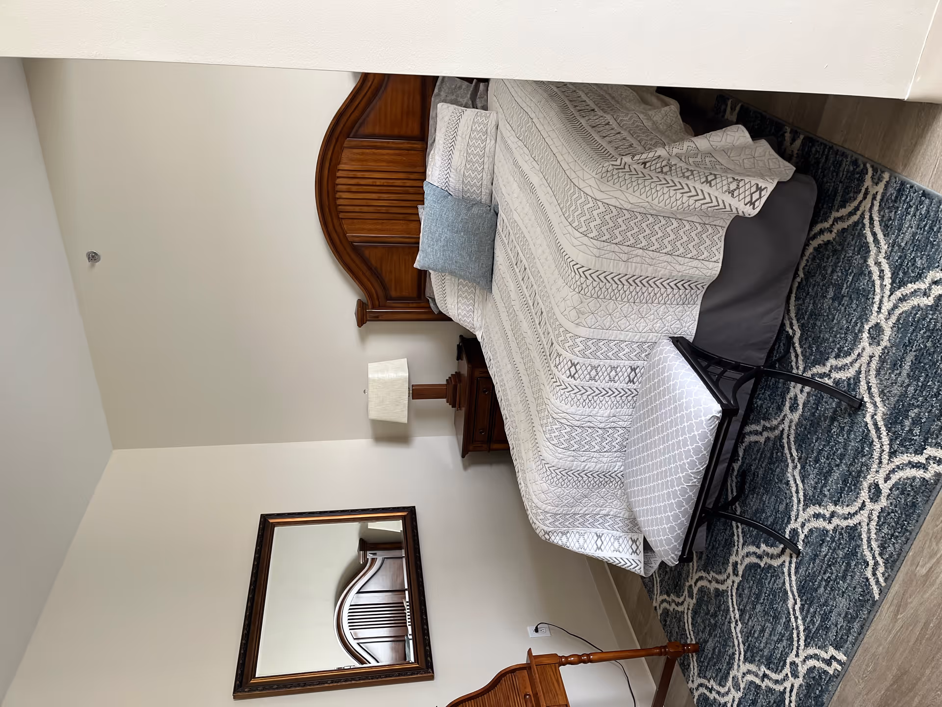 A furnished bedroom with a wooden headboard bed, patterned bedding, a nightstand with lamp, a small bench at the foot, and a patterned area rug.