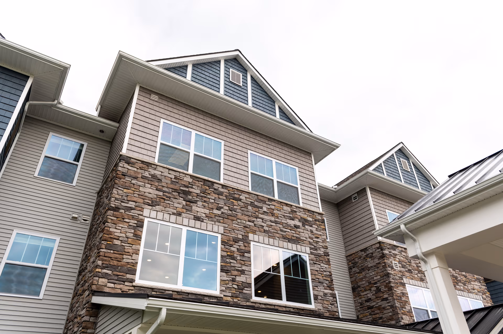 Upward view of a multi-story building with a combination of stone and siding exterior walls, multiple windows, and a metal roof section under an overcast sky.