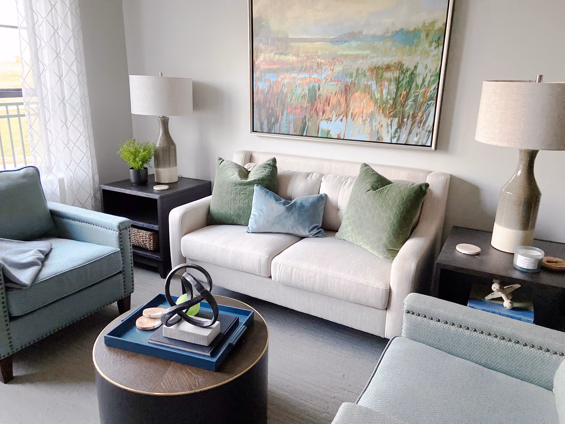 A cozy living room with a white sofa adorned with two green pillows and one blue pillow. There are two light blue armchairs with nailhead trim, a round wooden coffee table with decorative items, two dark side tables each with a lamp, a small plant, and a candle. A large colorful painting hangs on the wall above the sofa, and sheer white curtains cover a window letting in natural light.