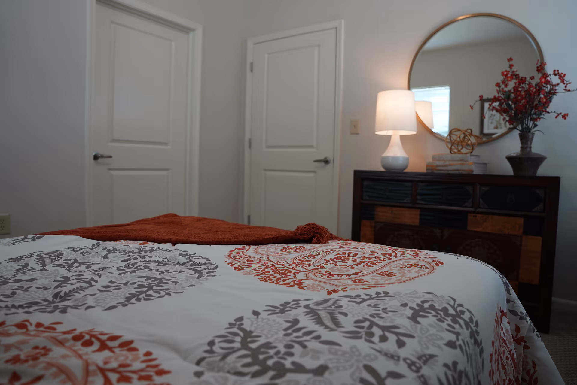 A bedroom with a bed covered in a white, gray, and orange patterned bedspread. There is a burnt orange throw blanket at the foot of the bed. In the background, there are two white doors, a dark wooden dresser with multiple drawers, a round mirror hanging above the dresser, a white table lamp, and a vase with red flowers.