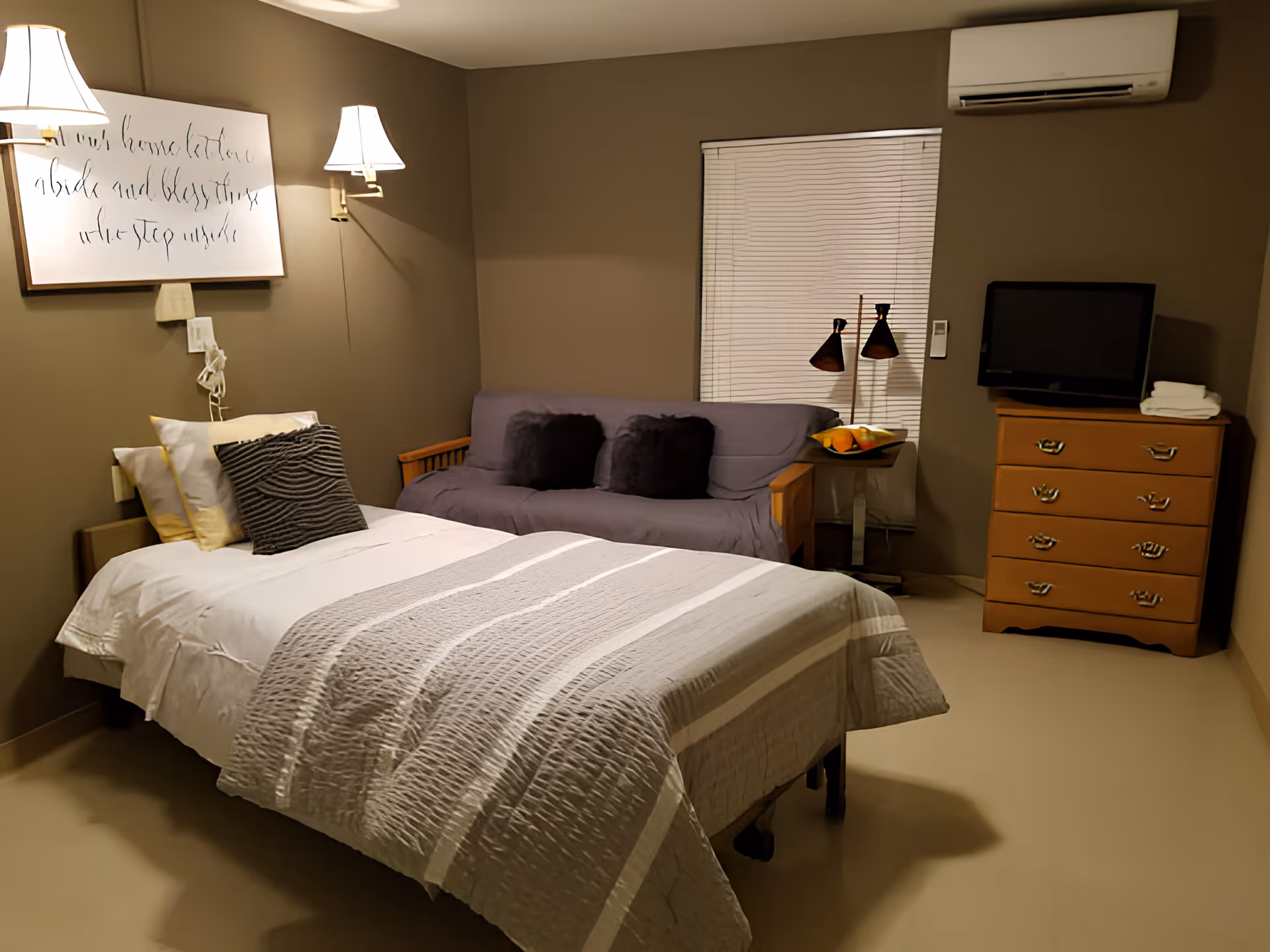 A cozy bedroom with a single bed covered in white and gray bedding, two pillows, and a striped cushion. On the wall above the bed are two wall-mounted lamps and a framed piece of calligraphy art. Next to the bed is a purple sofa with two dark cushions. A small side table with a fruit bowl and a dual-headed lamp is positioned near a window with closed blinds. A wooden dresser with a flat-screen TV and folded towels on top is against the opposite wall. An air conditioning unit is mounted above the window.